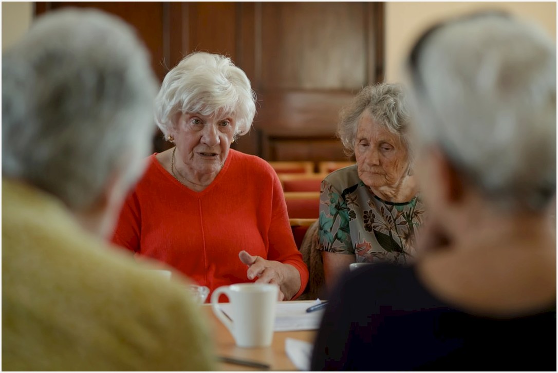 Elderly women sit around a table, talking.