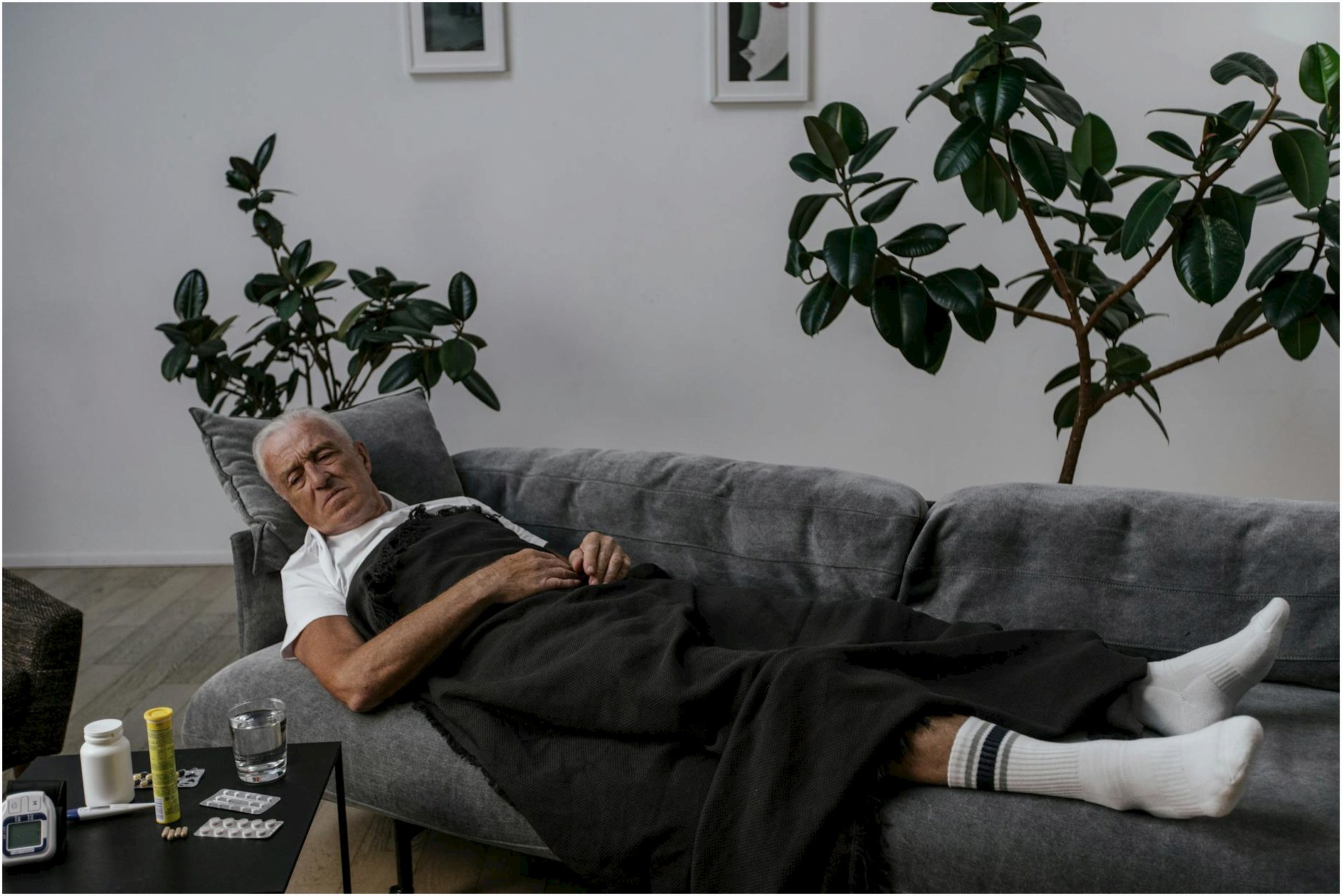 Elderly man resting on sofa with medical supplies nearby, surrounded by houseplants.