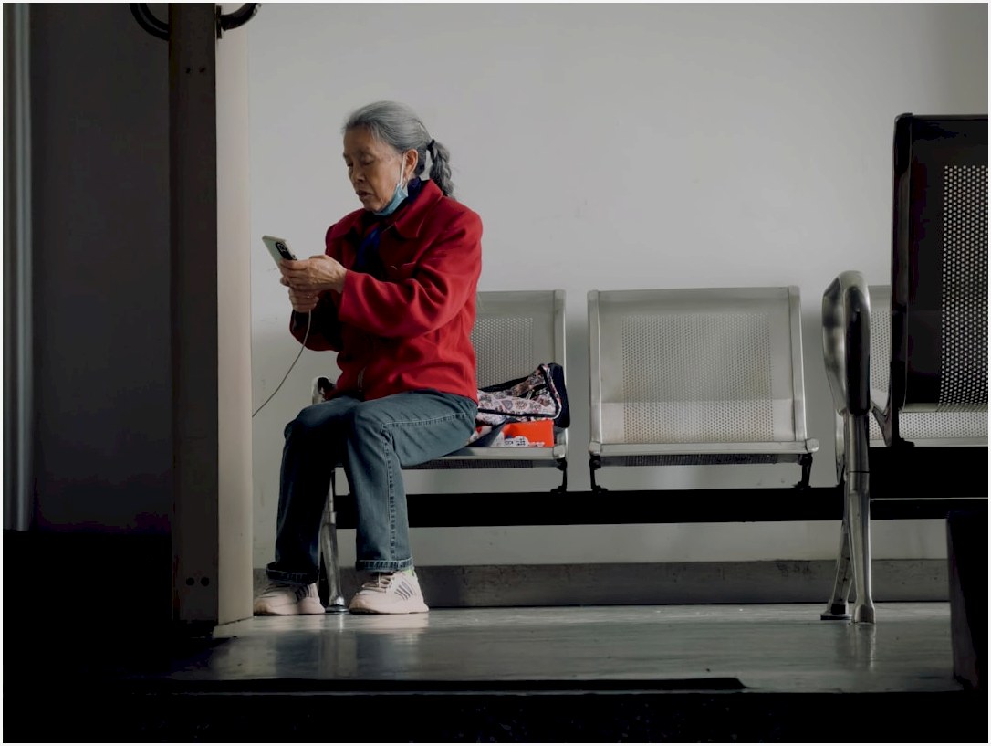 Elderly woman in red jacket uses smartphone while sitting.