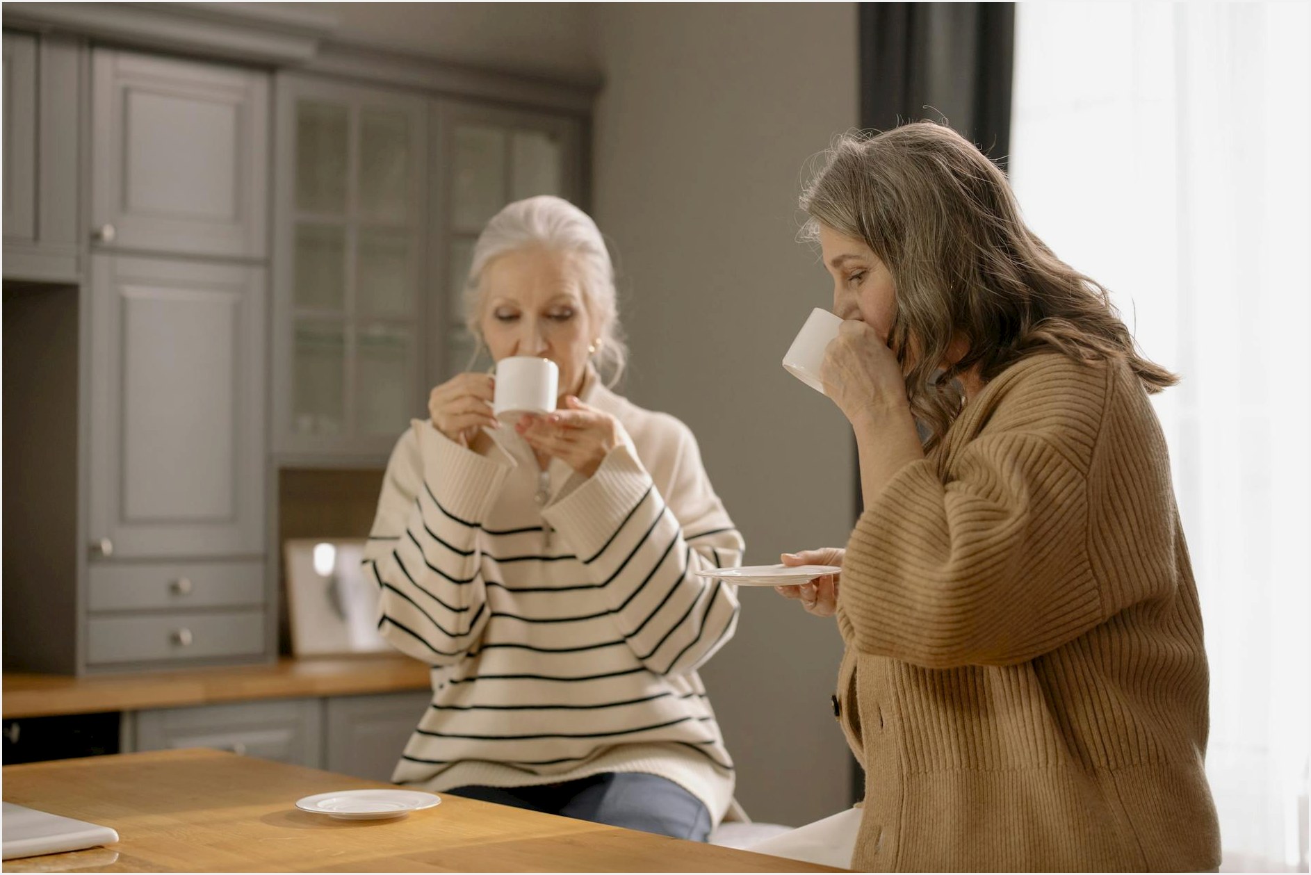 Two senior women relaxing and sipping coffee at a kitchen table, enjoying each other