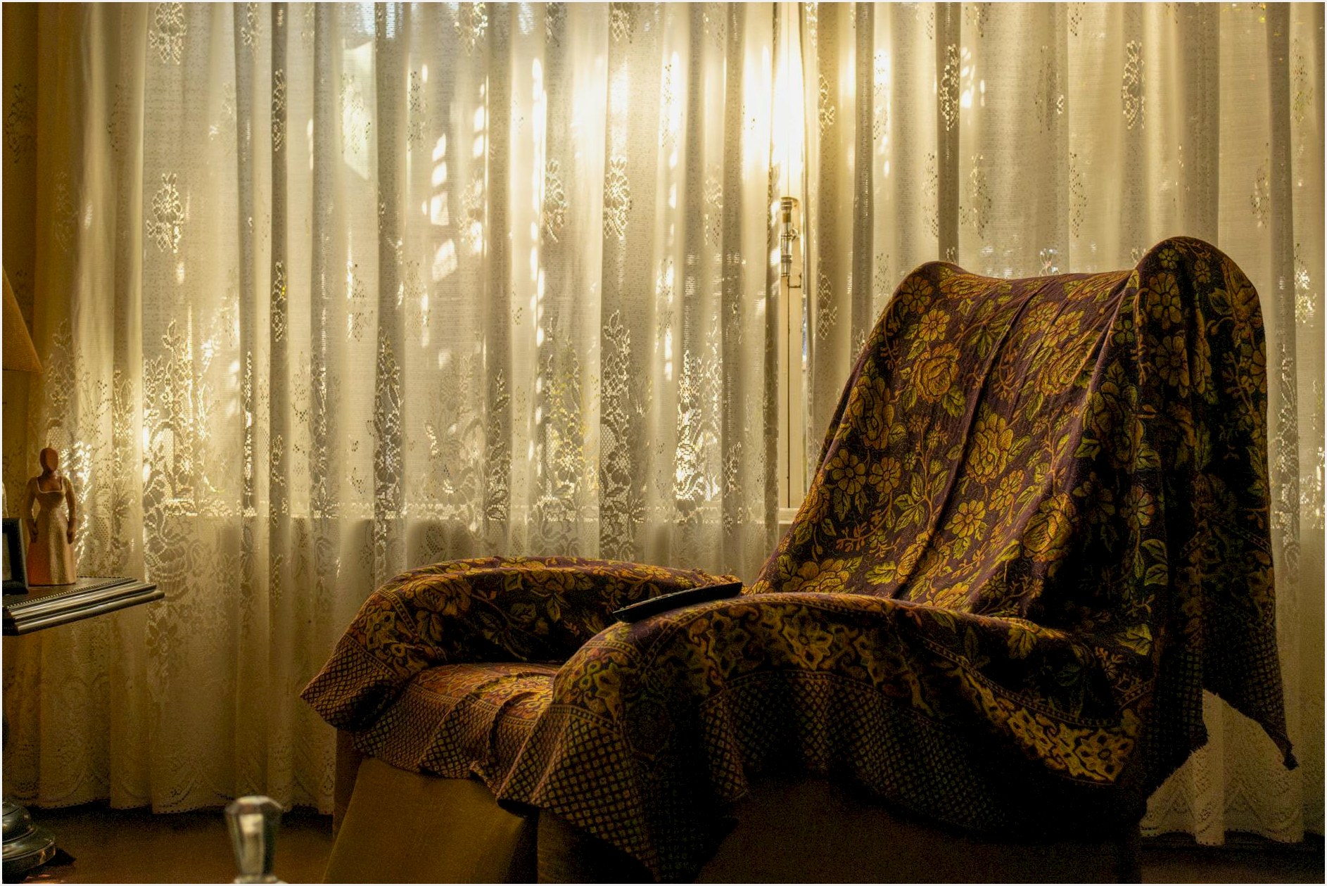 A cozy armchair draped with a vintage floral blanket near an elegant curtain-lit window.