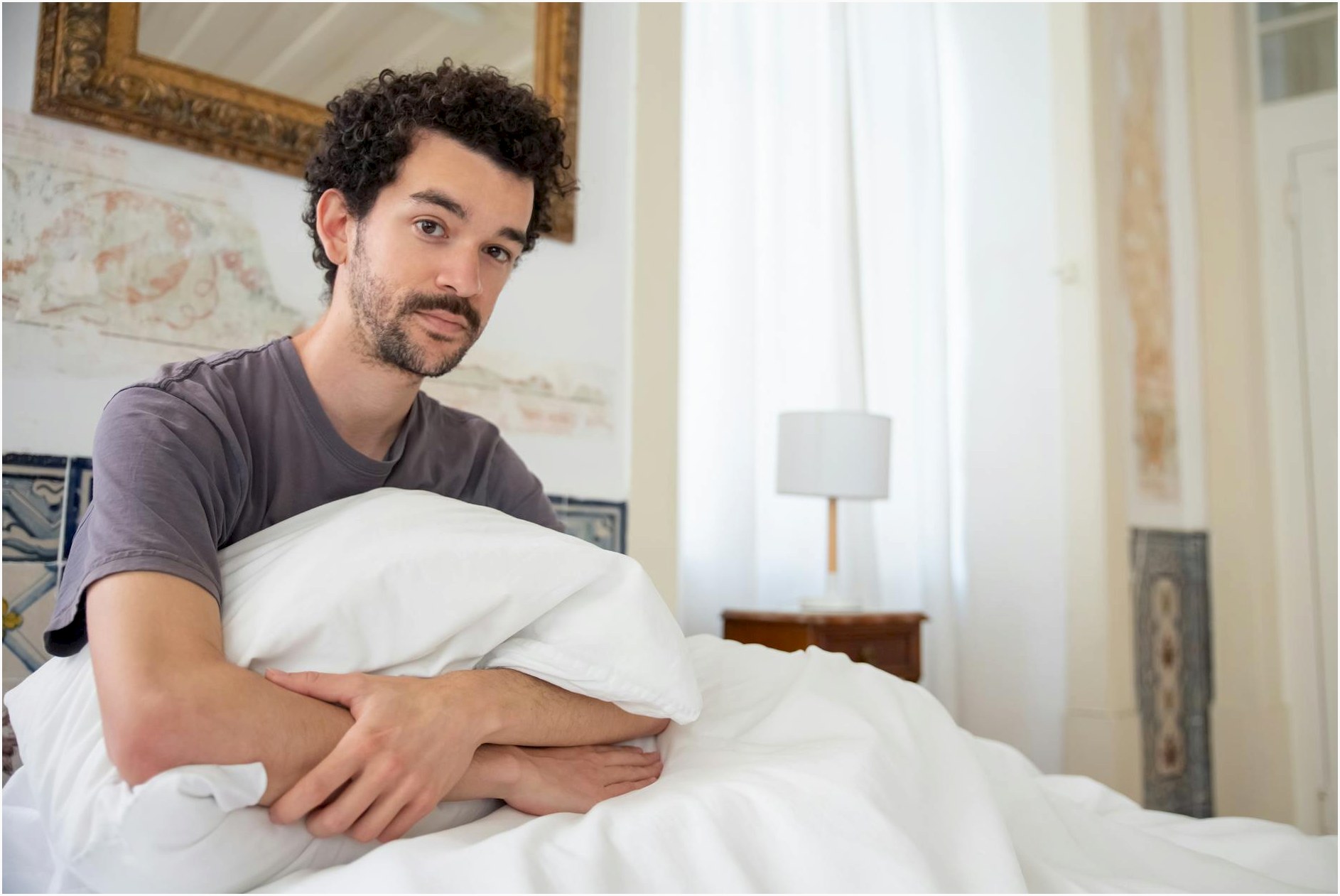 A man sits comfortably on a bed in a stylish Portuguese bedroom, holding a pillow.